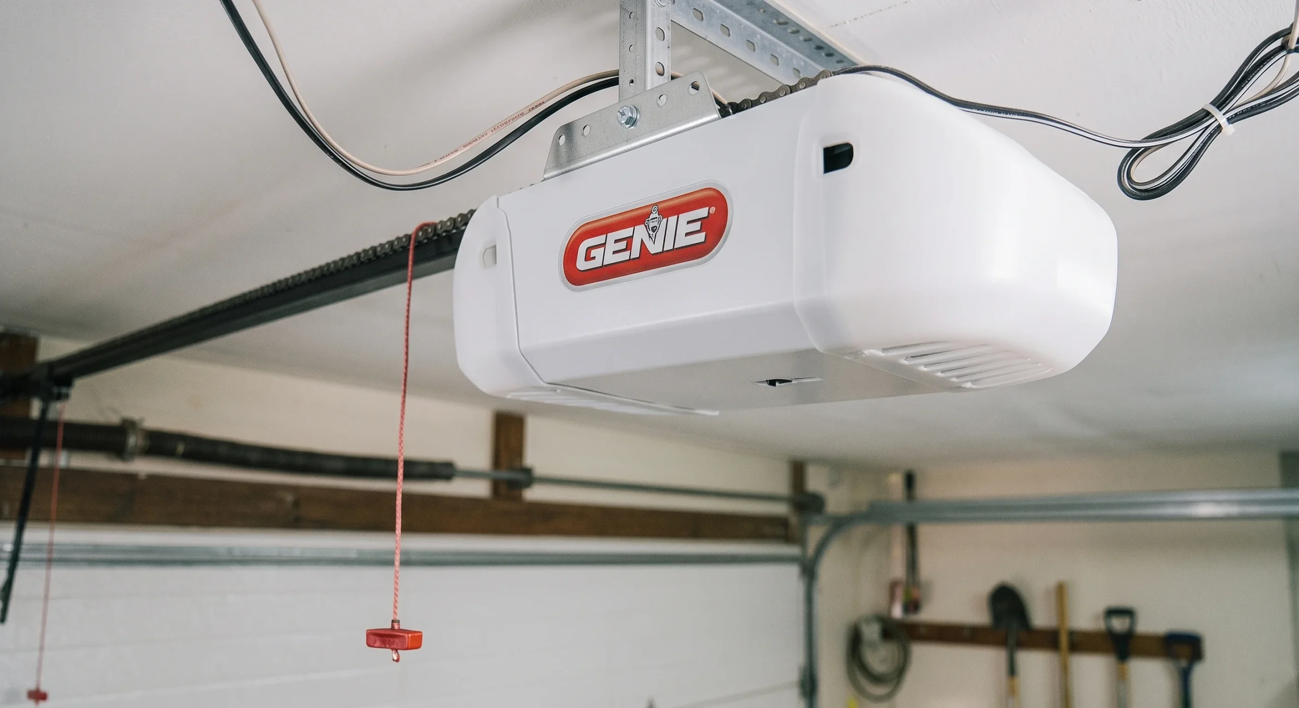 A white ceiling-mounted Genie motor unit with connected chain rail and manual release pull cord for Smart Garage Door Opener Maintenance.